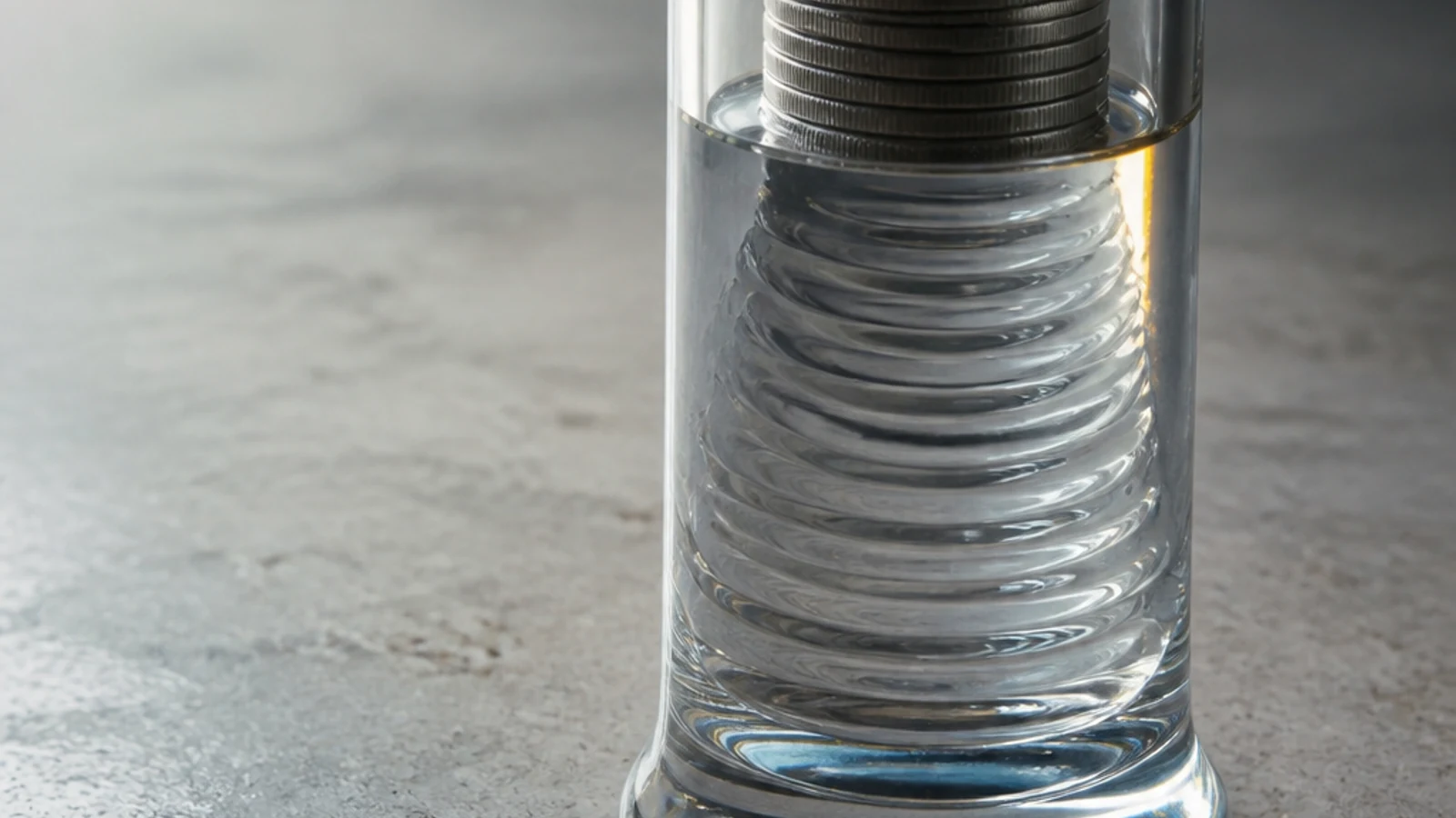 A clear glass measuring cylinder holds stacked coins that appear to dissolve into ripples on a pale surface.